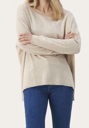 Kvinde iført beige langærmet sweater og blå jeans, stående med armene krydset mod en ensfarvet lys baggrund.