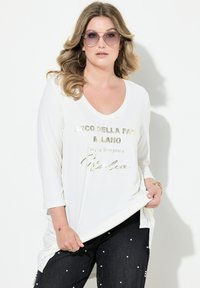 Weißes Langarmoberteil mit V-Ausschnitt und goldener Schrift. Kombiniert mit dunklen Jeans, die mit weißen Perlen akzentuiert sind.