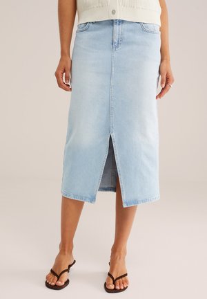 Ženska oblečena v svetlo modro jeans midi krilo z odprtim sprednjim razporkom, kombinirano z brezrokavnikom, kremne barve, iz pletenine in črnimi natikači.