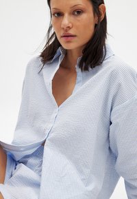 Light blue and white striped shirt with a button-down collar, featuring long sleeves and a relaxed fit. Textured fabric with vertical lines.