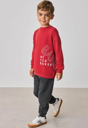 Băiat tânăr în picioare, purtând un tricou roșu cu mâneci lungi cu textul „Baker by Ted Baker”, pantaloni închiși la culoare și adidași albi cu tălpile de gumă.