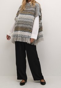 En person som bär en flerfärgad stickad poncho med fransar över en vit skjorta och svarta vida byxor tillsammans med svarta balettklackar.