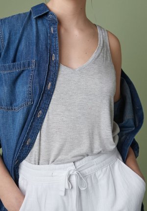 Denim skjorte over grå tank top, kombineret med hvide, snoretræk, linned bukser. Baggrunden er en dæmpet grøn. Funktioner inkluderer en brystlomme og knapdetaljer.