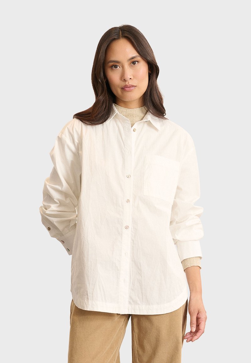 Witte oversized button-up shirt gemaakt van katoen met een borstzak, beige knopen en een licht gekreukt textuur, gecombineerd met lichtbruine broek.
