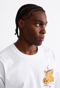 White cotton t-shirt with a round neckline featuring a graphic of a cartoon leopard wearing a cap, printed on the left chest.