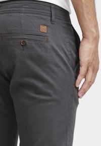 Gray trousers with a smooth texture, featuring a brown logo patch and a button closure on the back pocket. Simple design, tailored fit.