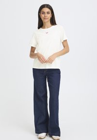 Krém színű, rövid ujjú pamut póló, kis rózsaszín szívvel a mellrészen. Széles szárú tengerészkék farmernadrággal párosítva.