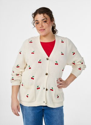 Frau mit lockigem Haar und Sommersprossen trägt eine cremefarbene Strickjacke mit Kirschmuster über einem roten Top, kombiniert mit blauen Jeans, steht mit der Hand in der Hüfte.