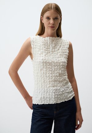 Top bianco senza maniche con un motivo testurizzato e ondulato. Presenta un collo alto e un orlo irregolare, abbinato a pantaloni in denim scuro.