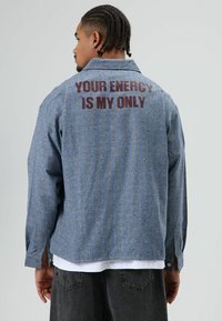 Blauw denim overhemd met een gestructureerd, gestreept patroon. Tekst "YOUR ENERGY IS MY ONLY" in donkerrood gedrukt op de achterkant. Lange mouwen, ontspannen pasvorm.