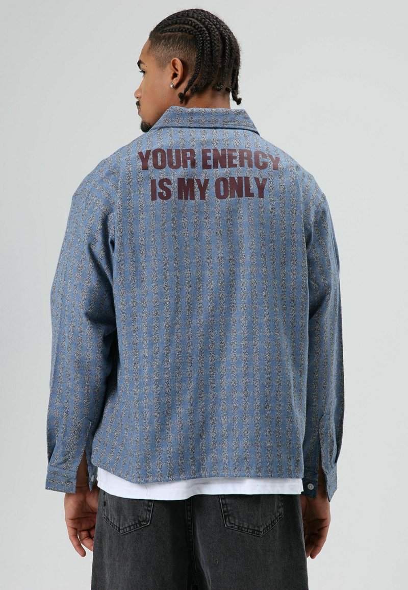Blauw denim overhemd met een gestructureerd, gestreept patroon. Tekst "YOUR ENERGY IS MY ONLY" in donkerrood gedrukt op de achterkant. Lange mouwen, ontspannen pasvorm.