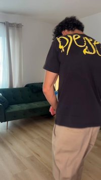 Person med krøllet hår iført en sort t-shirt med gul tekst og beige bukser, stående i en stue med en grøn sofa og gardiner.