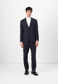 Traje navy con un corte ajustado; chaqueta de un solo pecho, solapas con muesca, dos bolsillos frontales, combinado con pantalones a juego y zapatos negros.