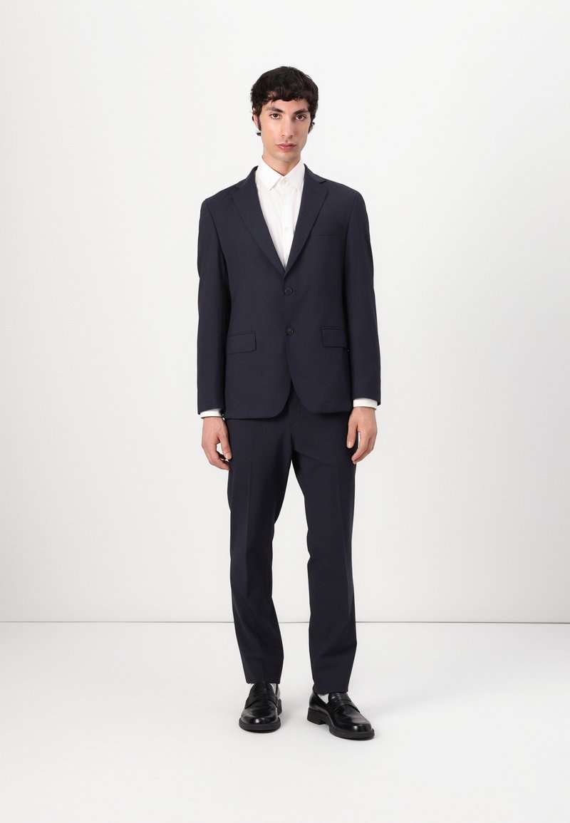 Traje navy con un corte ajustado; chaqueta de un solo pecho, solapas con muesca, dos bolsillos frontales, combinado con pantalones a juego y zapatos negros.