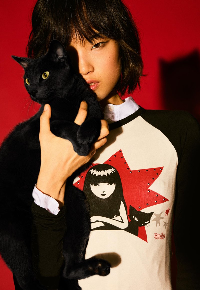 Duga crna i krem majica s dugim rukavima sa tiskanim dizajnom junaka i crvenim zvjezdastim detaljima, koju nosi osoba koja drži crnu mačku.