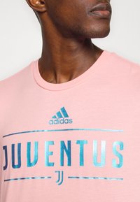 Světle růžové tričko s kulatým výstřihem, na kterém je modrý metalický logo "JUVENTUS" a nad ním modré logo Adidas.