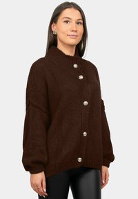 Pull marron tricoté avec un col montant, cinq grands boutons argentés et des manches volumineuses, présentant un tissu texturé.