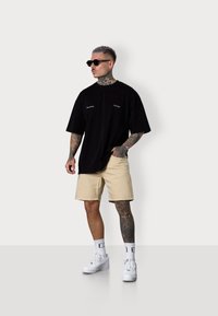 Tattooed man wears a black oversized t-shirt, beige shorts, and sunglasses, standing against a light gray background.
