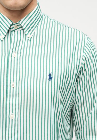 Άνδρας που φοράει πουκάμισο με κατακόρυφες πράσινες και λευκές ρίγες και ένα μπλε κεντημένο λογότυπο παίκτη πόλο στο στήθος.