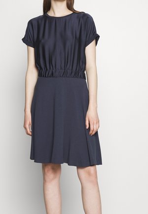 Woman wearing a navy blue dress with rolled short sleeves and gathered waistband, standing against a plain light background.