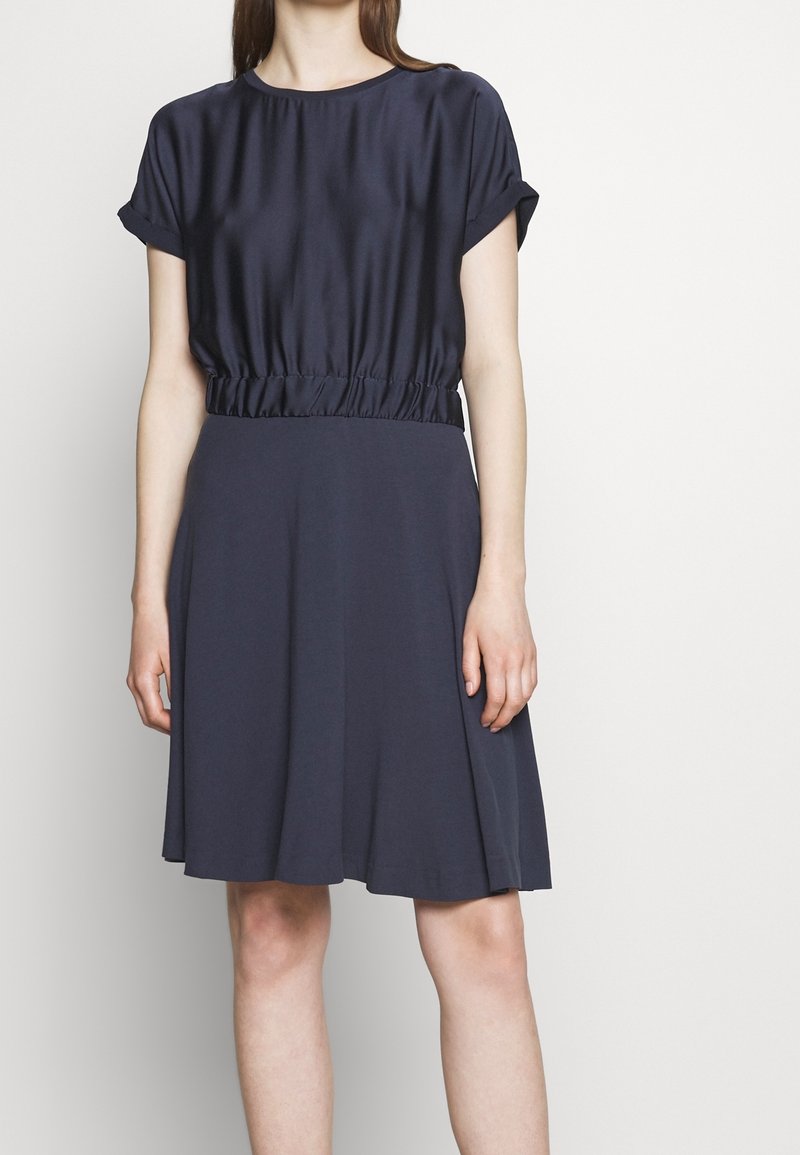 Femme portant une robe bleu marine avec des manches courtes retroussées et une ceinture froncée, debout devant un fond clair uni.