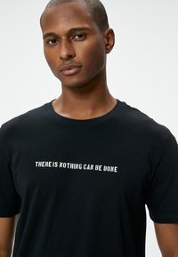 Sort bomulds T-shirt med korte ærmer og rund halsudskæring, med hvid tekst der står "DER KAN IKKE GØRES NOGET" på brystet.