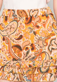 Saia floral multicolorida em laranja, com bainha ondulada e cintura elástica. O padrão inclui motivos paisley e flores.