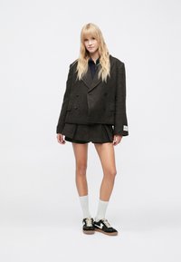 Barna kétsoros gyapjú blézer széles hajtókával, hozzá illő pliszírozott miniszoknyával. A modell fehér zoknit és fekete tornacipőt visel.