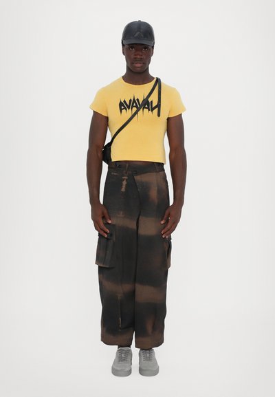 T-shirt giallo corto con testo grafico nero, abbinato a pantaloni cargo larghi neri e marroni con tasche e scarpe da ginnastica grigie.