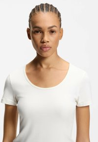 Ung person med fletninger iført en simpel hvid kortærmet skjorte, vendt fremad med et neutralt udtryk mod en hvid baggrund.