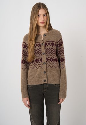 Femme aux longs cheveux blonds portant un cardigan marron boutonné avec des motifs géométriques bordeaux et un pantalon foncé, debout devant un fond uni.
