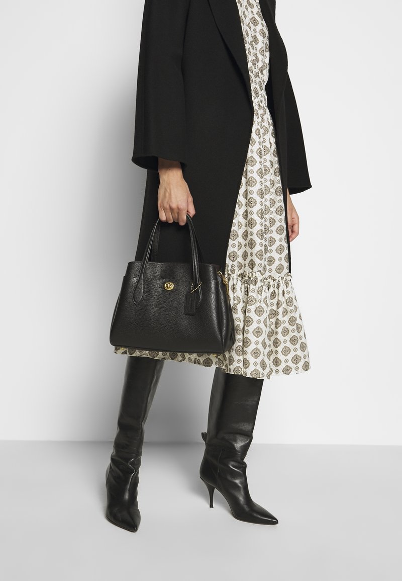 Personne portant un manteau noir et une robe à motifs tient un sac à main en cuir noir et porte des bottes noires à talons montant jusqu'aux genoux.