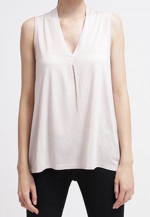 Beżowy, bezrękawnik z dekoltem w serek i plisą z przodu, noszony z czarnymi spodniami na stojącej osobie.