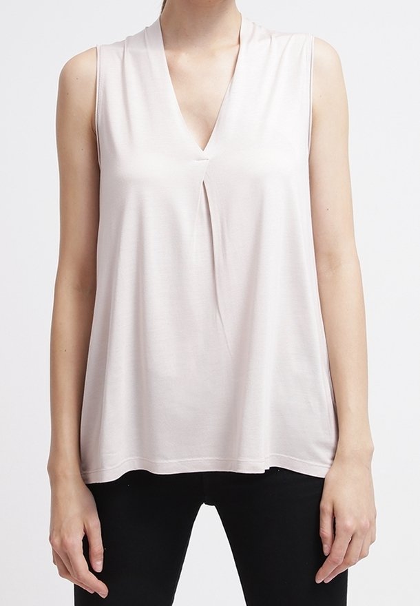Haut beige clair sans manches avec un col en V et un pli central à l'avant, porté avec un pantalon noir sur une personne debout.