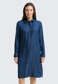 Robe en jean d'un bleu profond, avec un col pointu et un détail de nœud papillon. Fermeture à boutons sur le devant, manches longues et coupe décontractée.