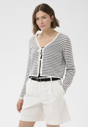 Kobieta ubrana w czarno-biały paskowany krótki kardigan na czarnej bluzce, białe szorty z paskiem, stoi z jedną ręką w kieszeni.
