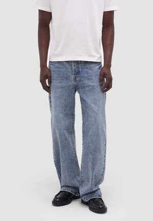 Uomo che indossa jeans larghi azzurri, scarpe nere e una maglia bianca a maniche corte, in piedi contro uno sfondo semplice.