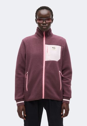 Personne portant une veste polaire violette avec des fermetures éclair roses et une poche poitrine rose clair, associée à un pantalon sombre et des lunettes de soleil foncées.
