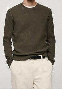 Uomo che indossa un maglione verde oliva a coste, pantaloni beige con una cintura nera, mani nelle tasche, su uno sfondo chiaro e semplice.