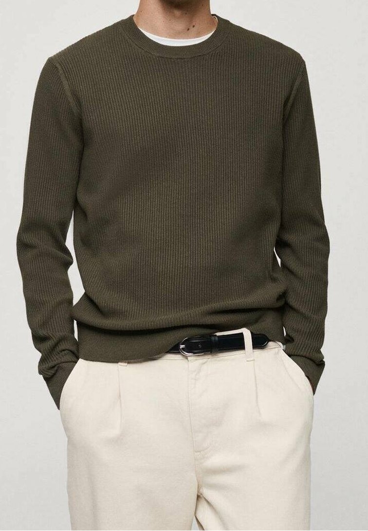Uomo che indossa un maglione verde oliva a coste, pantaloni beige con una cintura nera, mani nelle tasche, su uno sfondo chiaro e semplice.