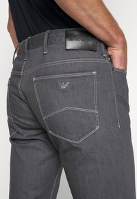 Man som bär mörkgrå Emporio Armani-jeans med synlig bakficka, logobadge och svart märkt läderpatch på midjan.