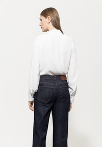 White blouse with long sleeves and button details, paired with dark denim jeans featuring a brown leather patch at the back.