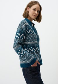 Gilet en tricot teal avec des motifs géométriques en blanc et marron clair. Il présente une fermeture éclair à l'avant, des poignets côtelé et une texture douce.