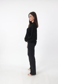 Maglione nero over-size con polsini e orlo a costine, abbinato a jeans neri aderenti e sandali con tacco beige. Sfondo bianco uniforme.