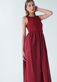 Dolga rdeča obleka z oprijetim zgornjim delom s smocked detajli in tekočo krilno linijo. Brezrokavnik, narejena iz lahkotne tkanine, gladke teksture z majhnimi nagubi.