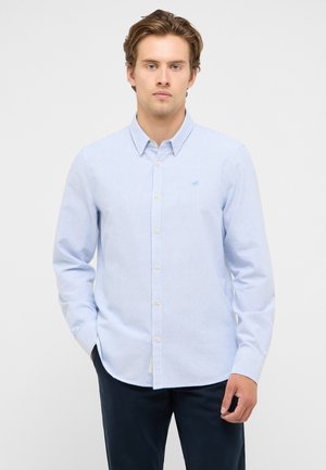 Young man with short hair wearing a light blue and white striped button-up shirt and dark pants standing against a plain background.