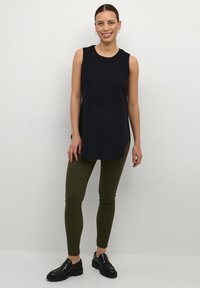 Top nero senza maniche con chiusura a bottoni, abbinato a leggings verdi oliva e mocassini neri. Texture liscia e design casual.