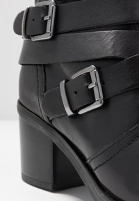 Black leather ankle boot featuring a thick block heel, adjustable silver buckles, and a smooth, textured finish with stitching details.