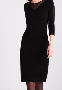 Musta mekko, jossa on istuva siluetti, jossa on V-pääntie ja kolme neljäsosaa hihat. Teksturoitu kuvio ja sileä kangas korostavat muotoilua.