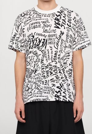 Witte T-shirt met korte mouwen met zwarte, handgeschreven stijl tekst over het hele shirt, waarbij de woorden "Versace Jeans Couture" herhaald worden in verschillende richtingen.